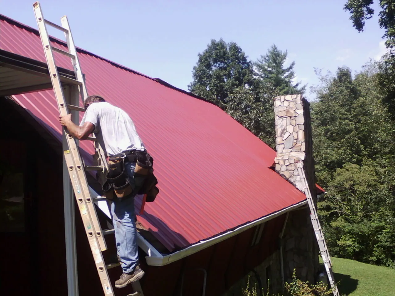 Roofer on a ladder beside a red metal roof and stone chimney — Soffit & Fascia Repair by Roofing Brothers in Leonardo